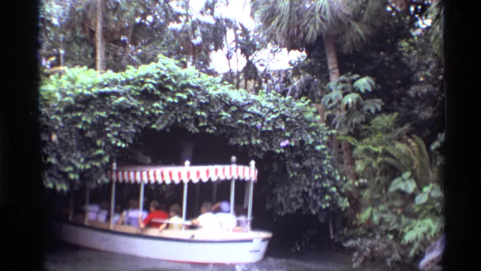 1974 Orlando home-movie frame of a small passenger boat under a striped canopy approaching a vegetation-covered tunnel.