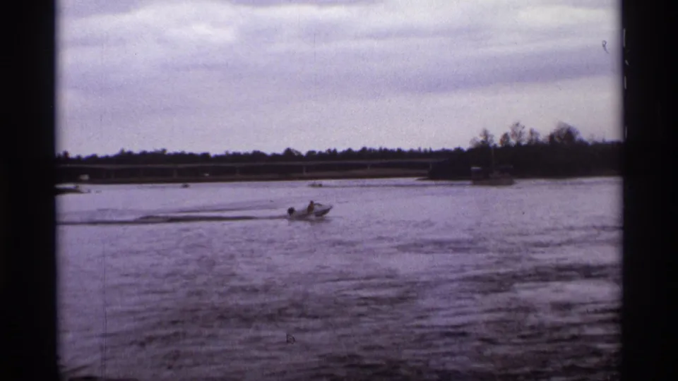 1974 Orlando home-movie frame of a small craft moving across a calm lagoon, with bridge and shoreline visible in the background.