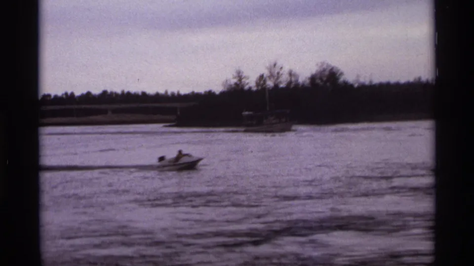 1974 Orlando lagoon footage showing a rider on a small motorized craft passing across frame with trees and a bridge in the distance.