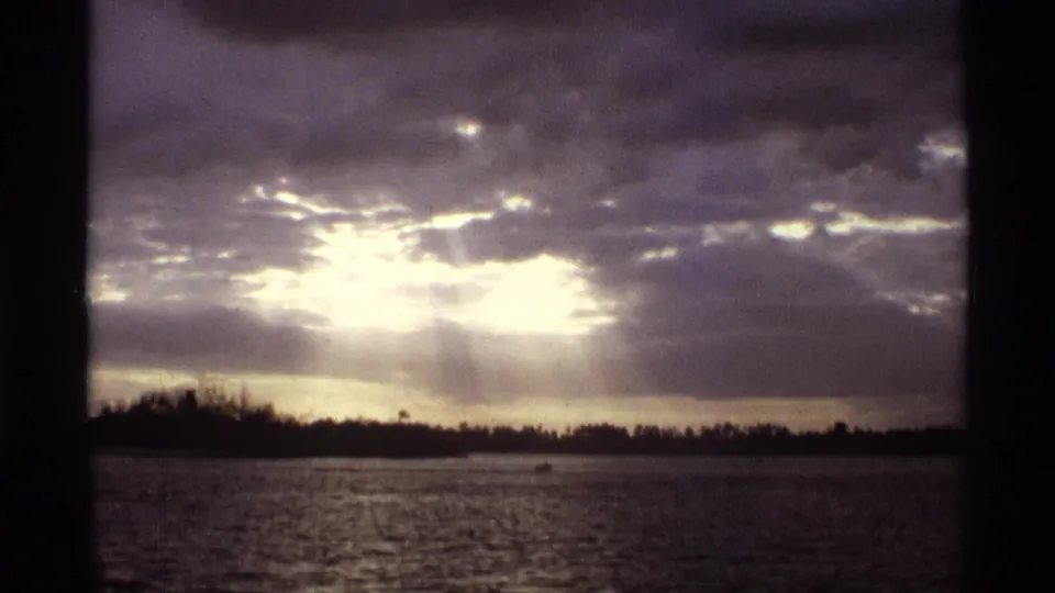 A sunset view over lake water shows bright sunbeams breaking through dark clouds above a low Florida shoreline.