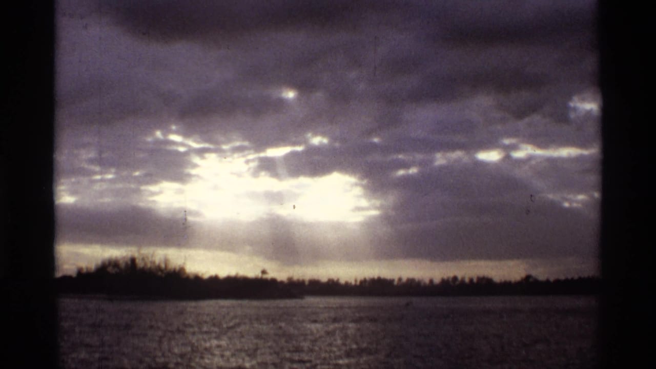 Sunlight breaks through heavy clouds over rippled water and a dark tree-lined shore.