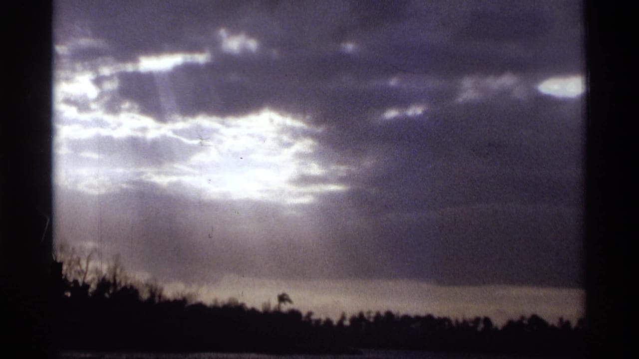 1974 Orlando-area 8mm frame of a bright break in storm clouds above a dark, tree-lined water horizon.