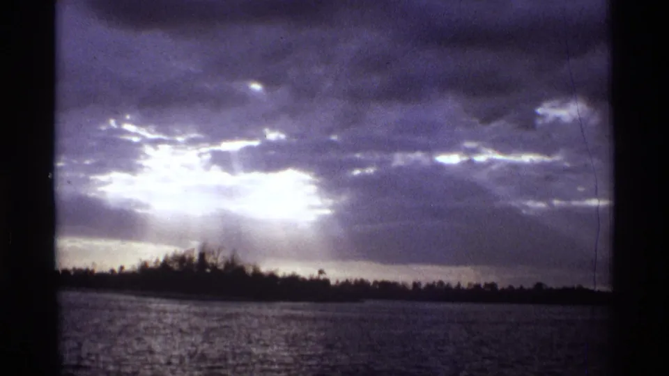 1974 Orlando-area 8mm frame with storm clouds, bright sun rays, rippled water, and a dark shoreline silhouette.
