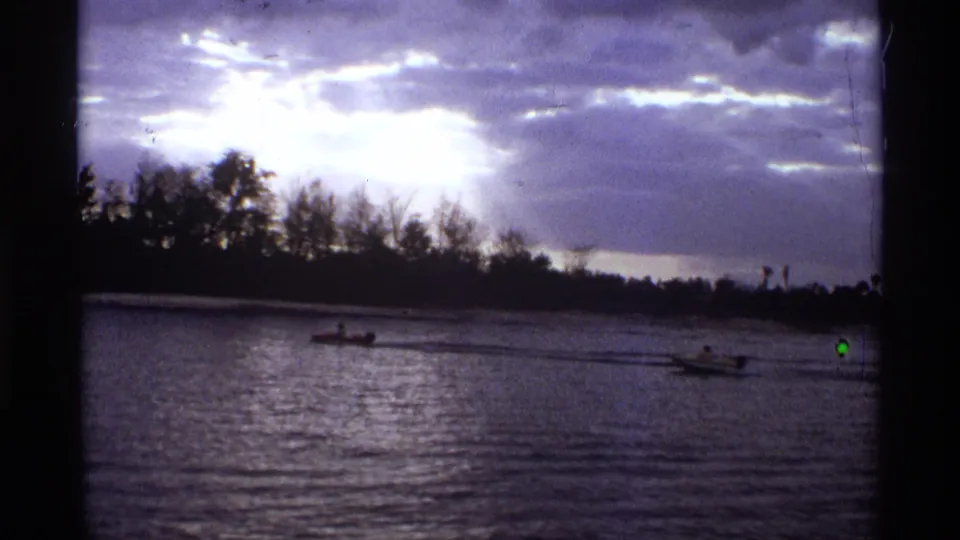 1974 Orlando-area 8mm frame showing two distant motorboats moving across rippled river water under bright clouds and a silhouetted shoreline.