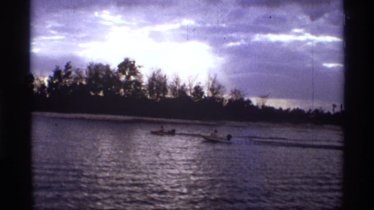 1974 Orlando-area home-movie frame with two small boats on a river, distant riders, dark trees, and a bright cloud break overhead.