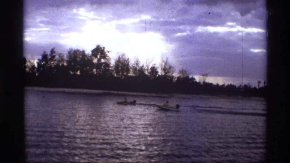 1974 Orlando-area home-movie frame with two small boats on a river, distant riders, dark trees, and a bright cloud break overhead.