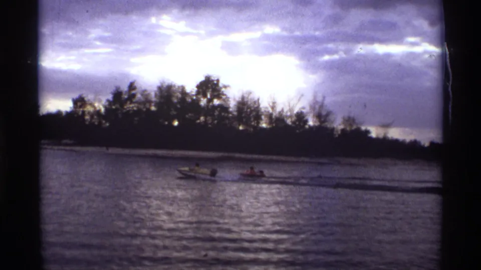 1974 Orlando-area 8mm frame showing two small motorboats near midriver, with riders visible at a distance and a dark tree line under purple clouds.