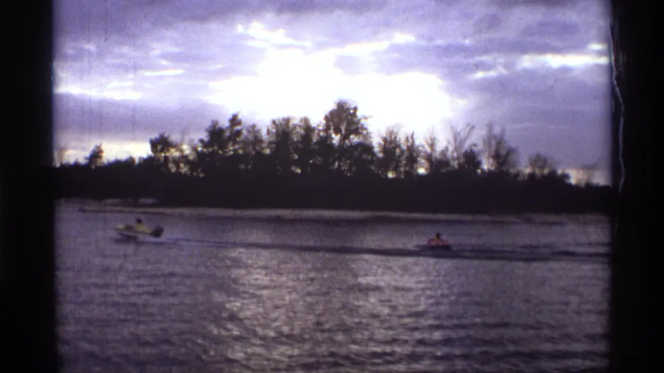 1974 Orlando-area home-movie frame with separated motorboats on a rippled river, distant riders, silhouetted trees, and bright clouded sky.