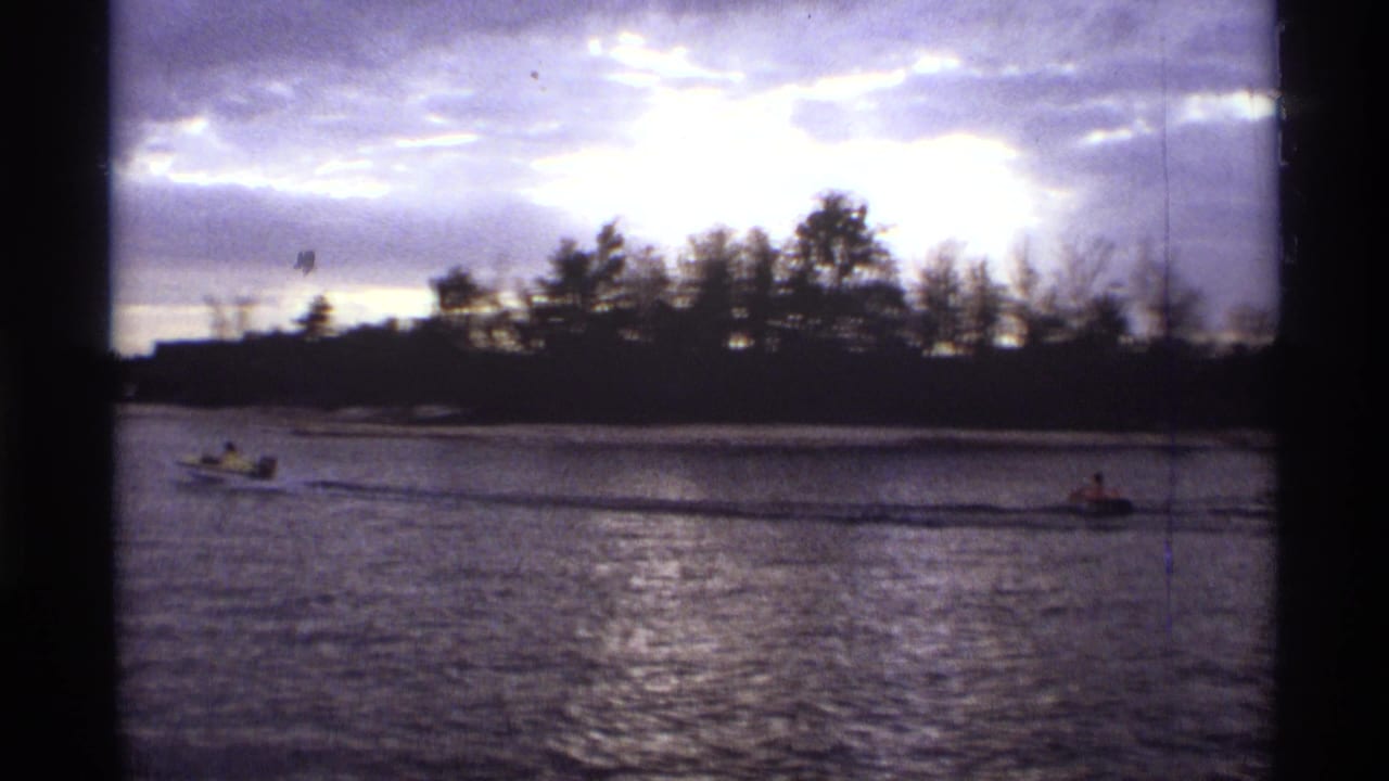 Small boat crosses a dark waterway below a cloudy sky and tree-lined shore.