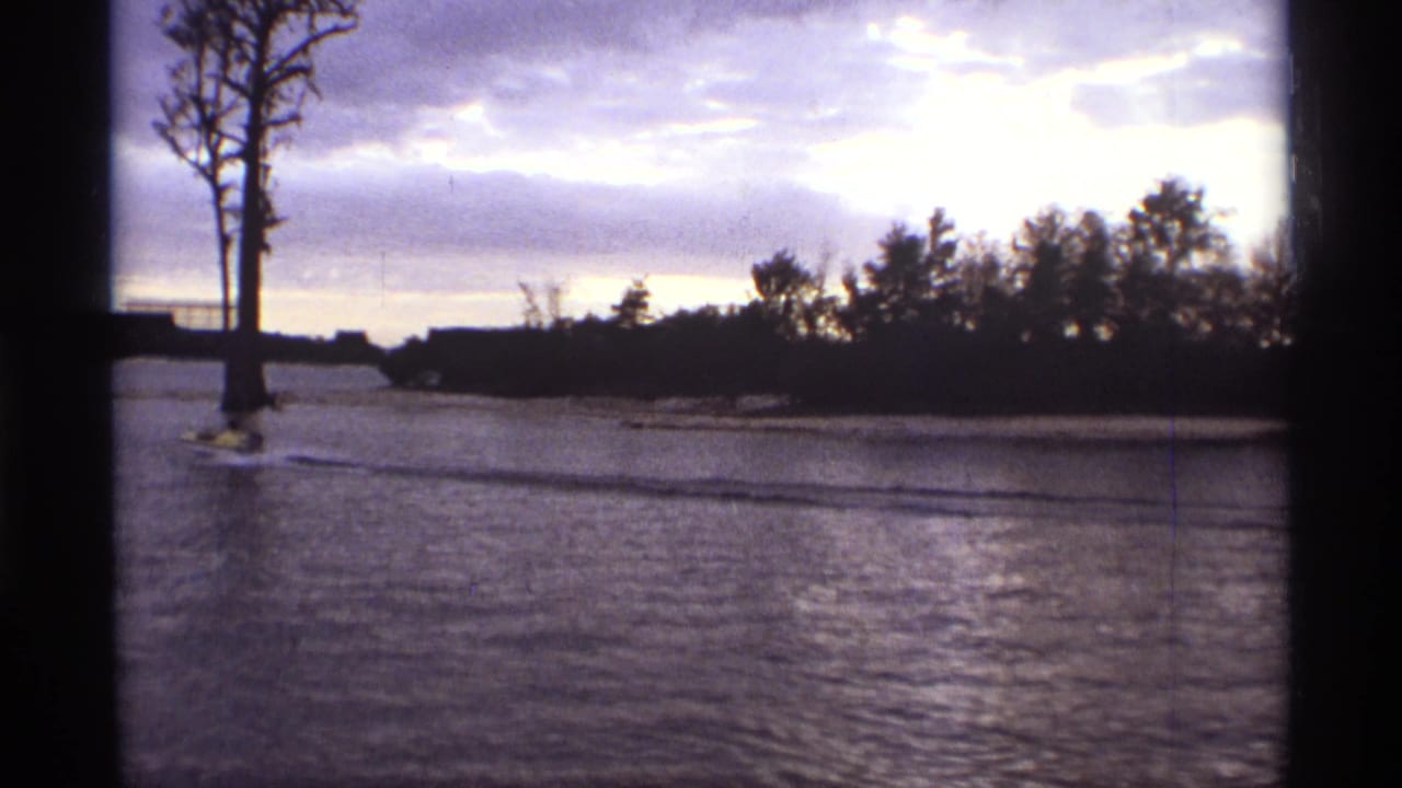 1974 Orlando home-movie frame with a small speedboat moving past a bare lakeside tree and low distant buildings.
