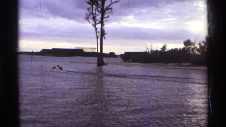 1974 Orlando 8mm frame showing a small speedboat near a tall bare tree, with low buildings and water in view.