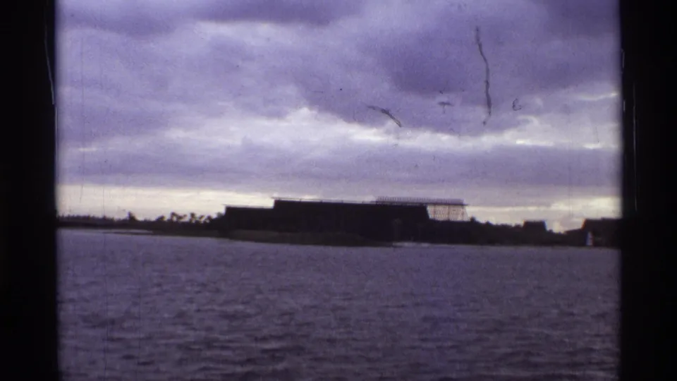 1974 Orlando home-movie frame with a broad water foreground, silhouetted building, and dense overcast clouds.