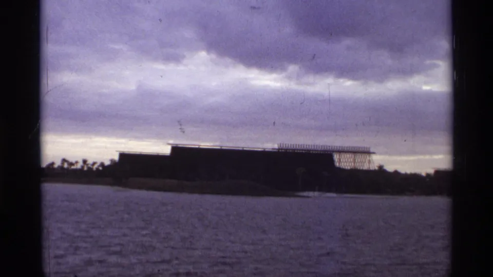 1974 Orlando 8mm frame with a dark low building near the shoreline, open water, and layered stormlike clouds.