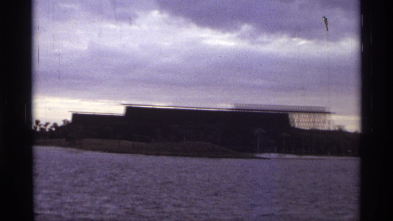 Wide view across water toward a low brown building under a cloudy sky.
