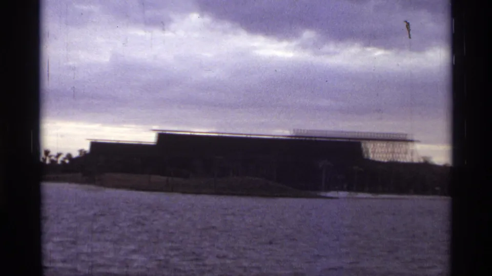 1974 Orlando 8mm frame showing a distant brown waterfront building, grassy bank and cloudy sky.