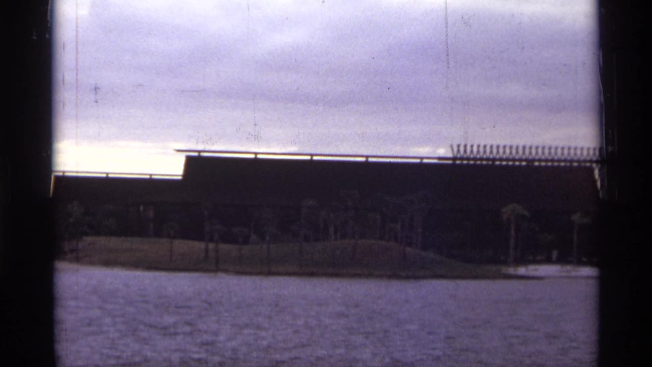 1974 Orlando home-movie frame with the brown building closer, palm trees along the bank and water in front.