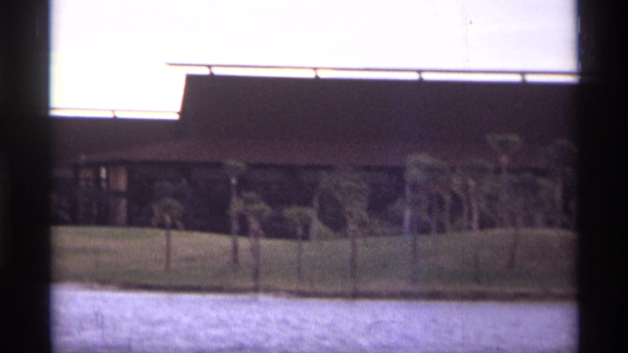 1974 Orlando home-movie frame showing a low modernist building beyond water, with palms, lawn, cloudy sky, and dark film borders.
