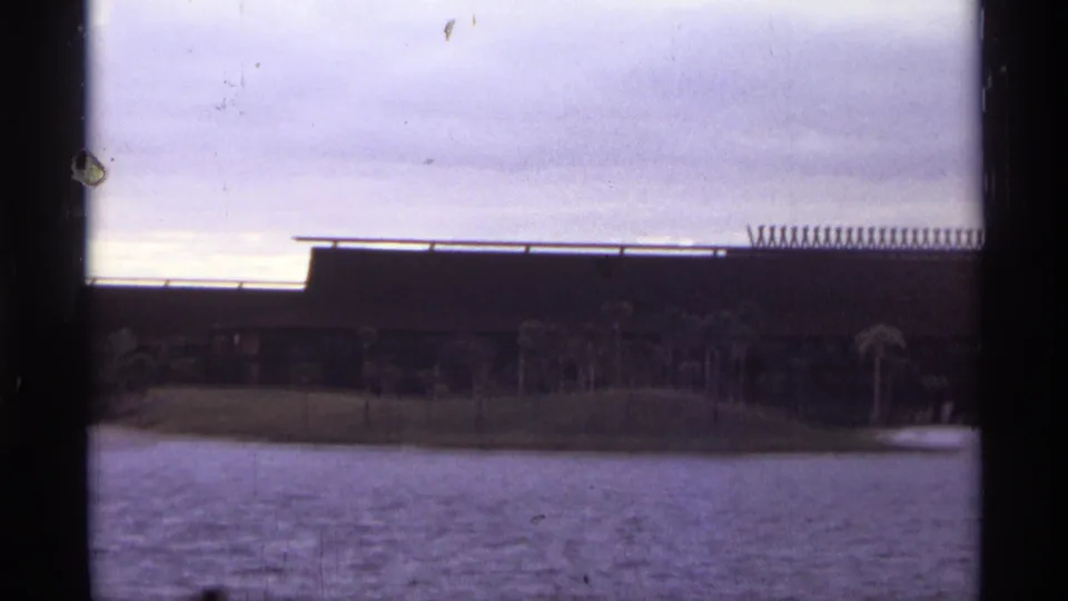 A faded 1974 Orlando 8mm frame shows water in the foreground, palms along the shore, and a low modernist building beneath clouds.