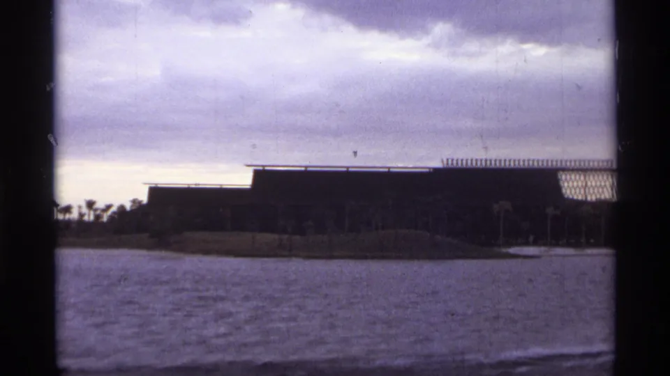 Wide 1974 Orlando home-movie view of rippled water, a palm-dotted shore, and a long low building with patterned facade detail.