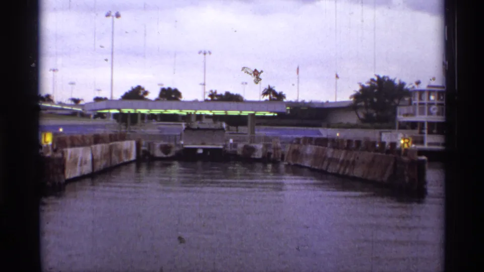 1974 Orlando-area 8mm frame showing a ferry-style dock approach with concrete walls, covered boarding area, flags, and palm trees.