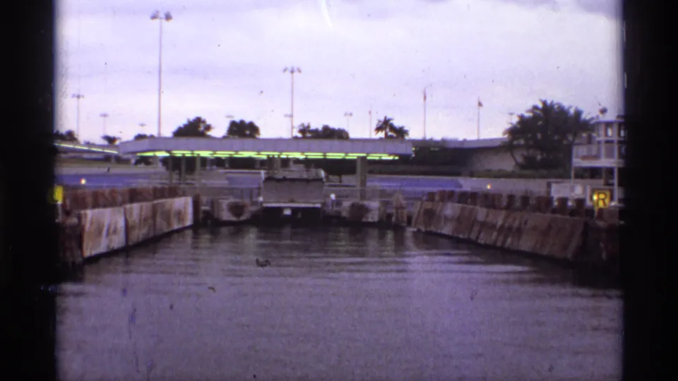 1974 Orlando-area dock channel with concrete sides, covered boarding structure, pole lights, flags, and palm trees in a faded 8mm frame.