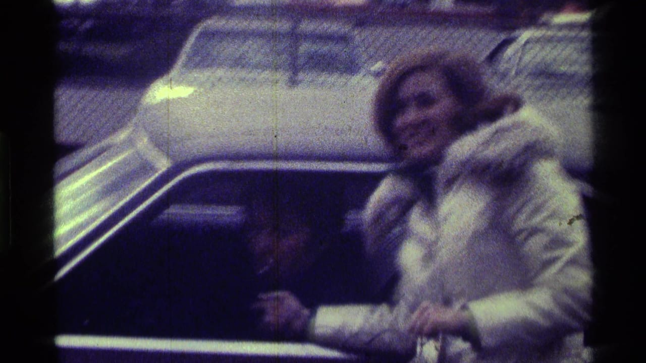 In 1976 Kentucky 8mm footage, a woman in a white fur-trimmed coat stands by a car with a microphone as a seated man is partly visible.