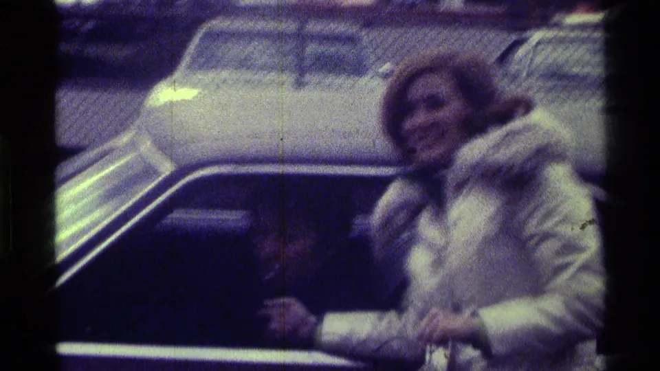 In 1976 Kentucky 8mm footage, a woman in a white fur-trimmed coat stands by a car with a microphone as a seated man is partly visible.