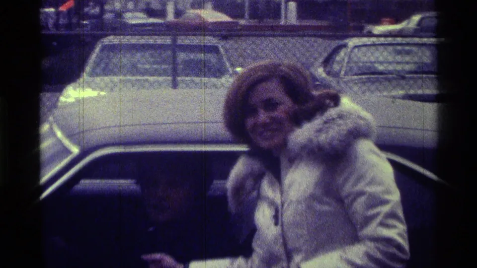 The woman smiles while standing beside the car, her microphone lowered, with the seated man dimly visible inside.
