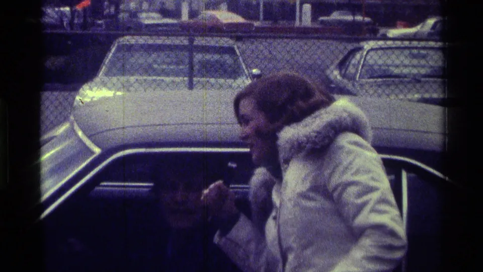 The woman leans in with the microphone toward the man inside the car, framed by parked vehicles and chain-link fencing.