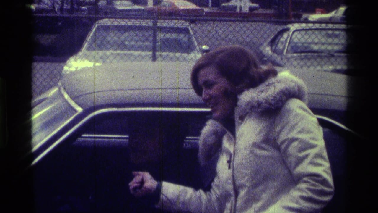 In 1976 Kentucky 8mm footage, a woman in a white winter coat leans toward a car window while holding a microphone.