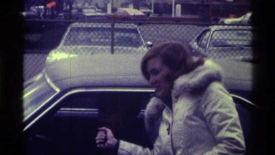 The interviewer in a white coat smiles and leans toward a person seated in a car, with parked vehicles and fencing behind her.