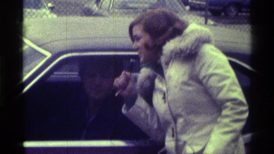 A white-coated interviewer holds a microphone near a man seated in a car, with 1970s parked cars visible in the background.