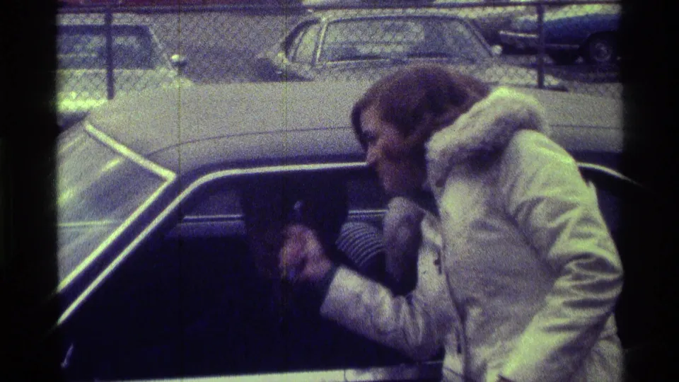 The woman continues the car-side interview, leaning forward with the microphone as parked cars and a fence fill the background.