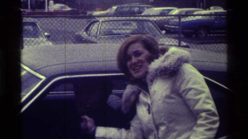 The woman smiles beside the car window, with chain-link fencing and 1970s-era parked cars visible in the background.