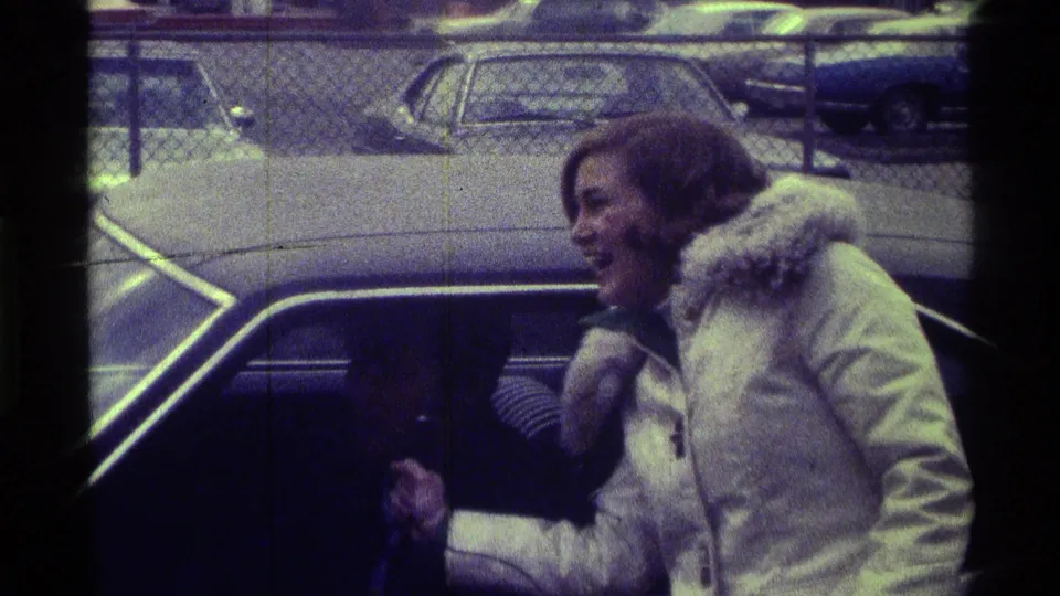 The woman laughs broadly and reaches toward the car window, framed by grainy 8mm footage and parked cars behind a fence.