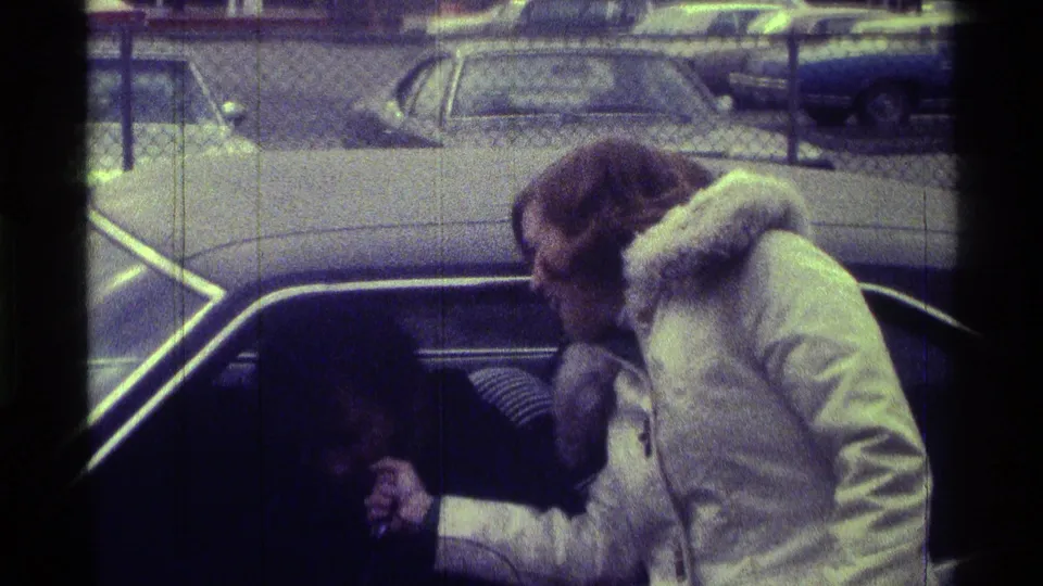 The woman turns toward the dark car interior, continuing the informal exchange in the Kentucky parking-lot home movie scene.