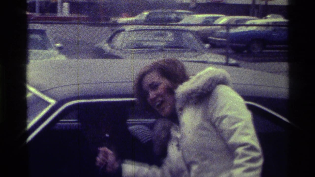 Woman in a white coat leans toward a car window in a parking lot.