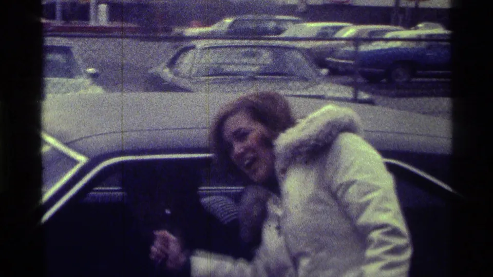 In 1976 Kentucky home-movie footage, a woman in a white fur-trimmed coat leans toward a dark car window in a lot with parked 1970s cars.