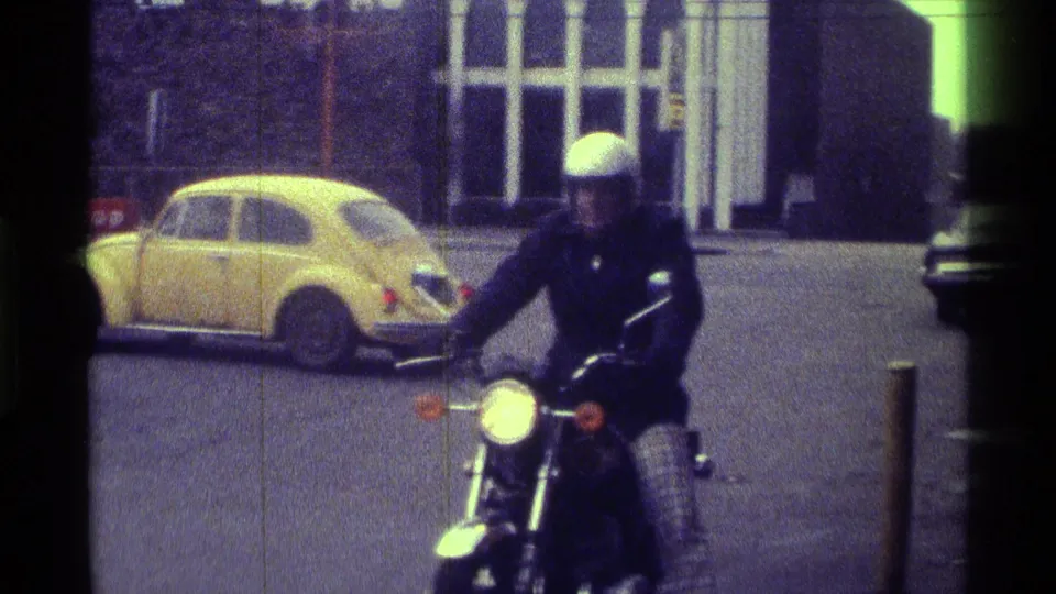 A helmeted motorcyclist in dark clothing rides through the 1976 Kentucky street scene with a yellow compact car and building windows behind him.