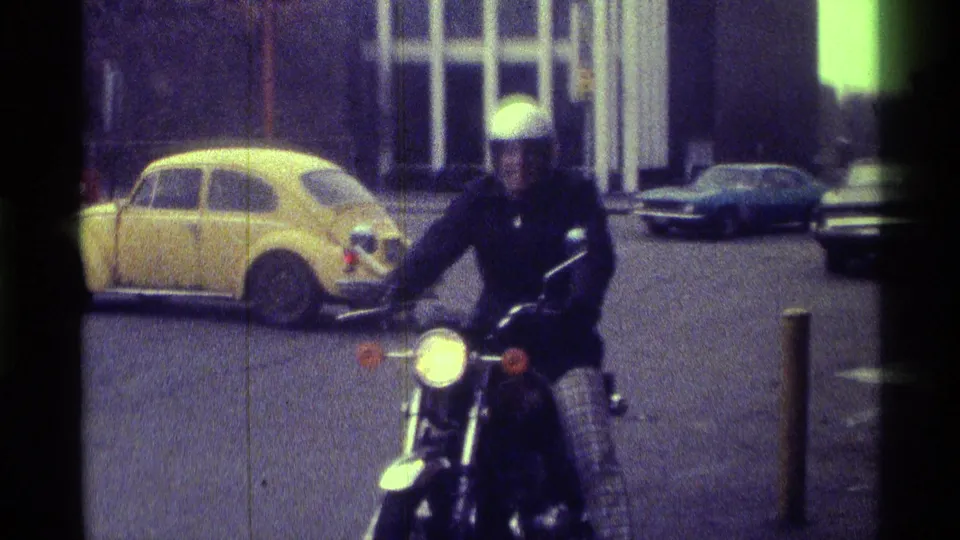 The motorcyclist moves closer to camera in the 1976 Kentucky footage, with a yellow compact car, bollards, and parked vehicles in view.
