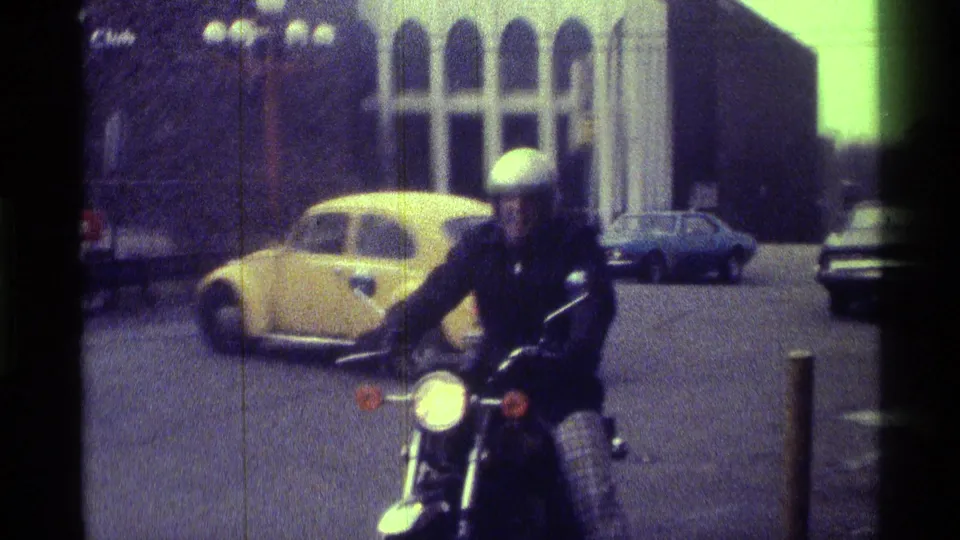 A helmeted rider passes through the 1976 Kentucky parking-area scene, framed by a yellow compact car, other parked cars, and tall building windows.