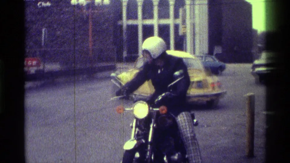 In Kentucky 1976 home-movie footage, a helmeted motorcyclist approaches or pauses on a city street near a yellow Volkswagen Beetle.