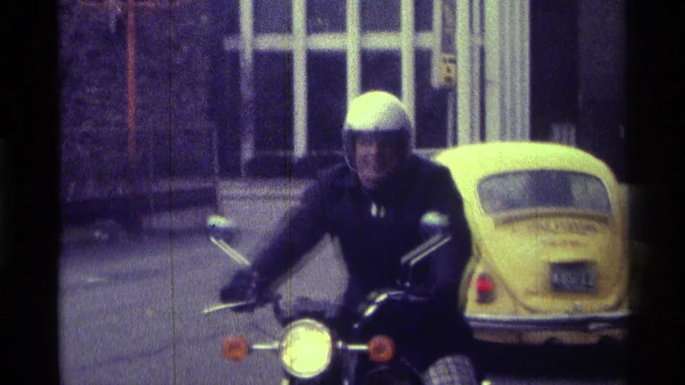 The rider faces the home-movie camera from his motorcycle, with a yellow Volkswagen Beetle and arched building windows in the background.