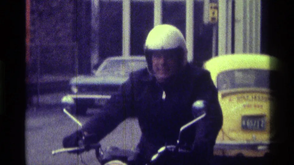 The motorcyclist appears to smile toward the camera on a Kentucky street, with parked cars and the yellow Volkswagen Beetle behind him.