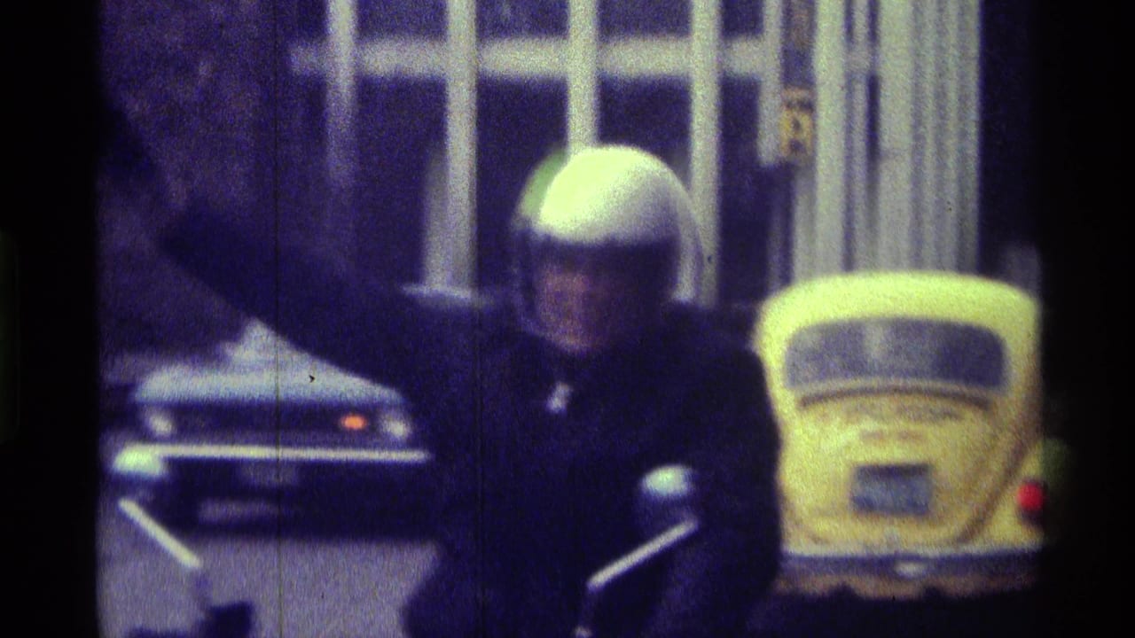 Helmeted motorcyclist waves on a street with a yellow Volkswagen Beetle behind him.