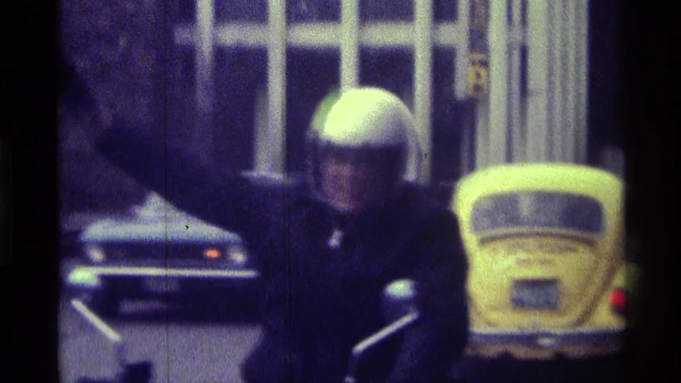 In 1976 Kentucky footage, a man in a white motorcycle helmet rides toward the camera while waving, with a yellow Volkswagen Beetle in the background.