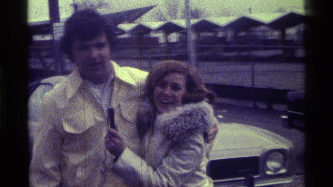 In 1976 Kentucky home-movie footage, a woman in a white fur-trimmed coat leans toward a man in a yellow jacket beside a light-colored car.