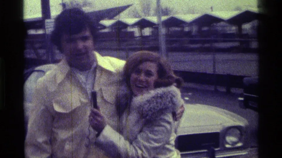 In 1976 Kentucky home-movie footage, a woman in a white fur-trimmed coat leans toward a man in a yellow jacket beside a light-colored car.