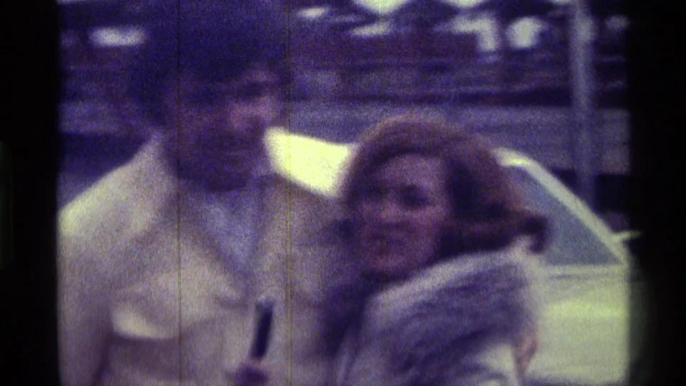 A closer 8mm frame shows the man and woman beside the car, with the woman in a white fur-trimmed coat and the man in a pale yellow jacket.