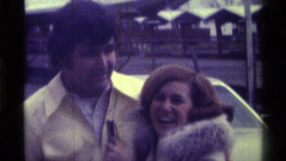 The woman smiles beside the man near the car, with soft 8mm color and outdoor structures visible behind them in Kentucky footage from 1976.
